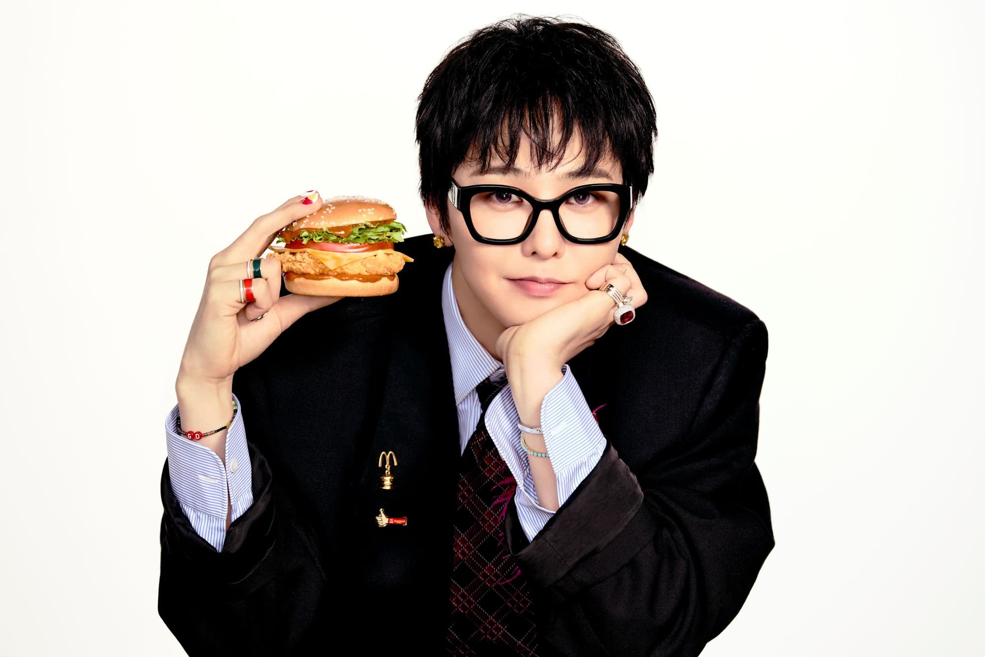 G-Dragon in glasses and a black blazer holding a McDonald’s chicken sandwich toward the camera