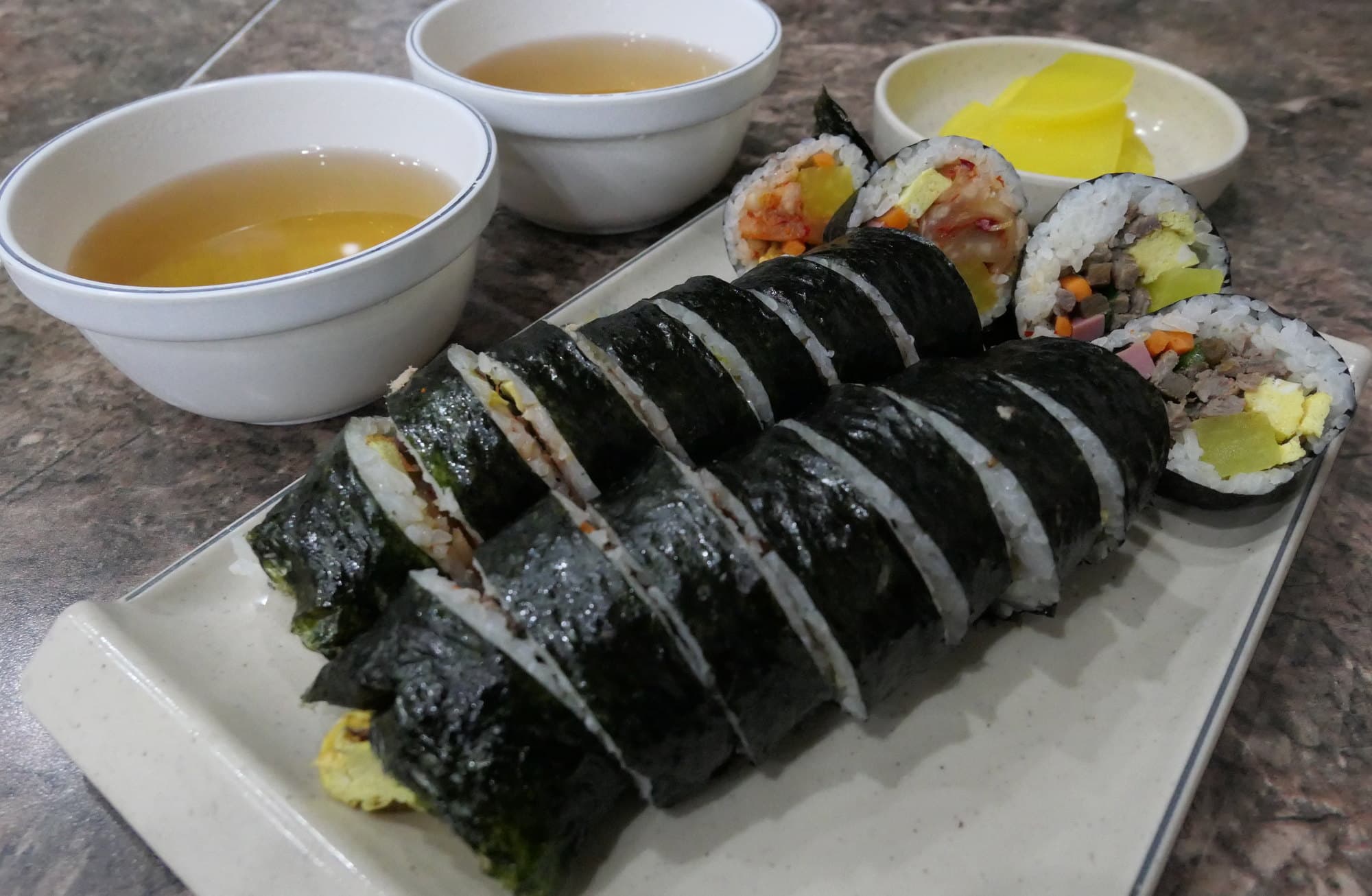 Sliced gimbap rolls with danmuji and broth cups on a stone table