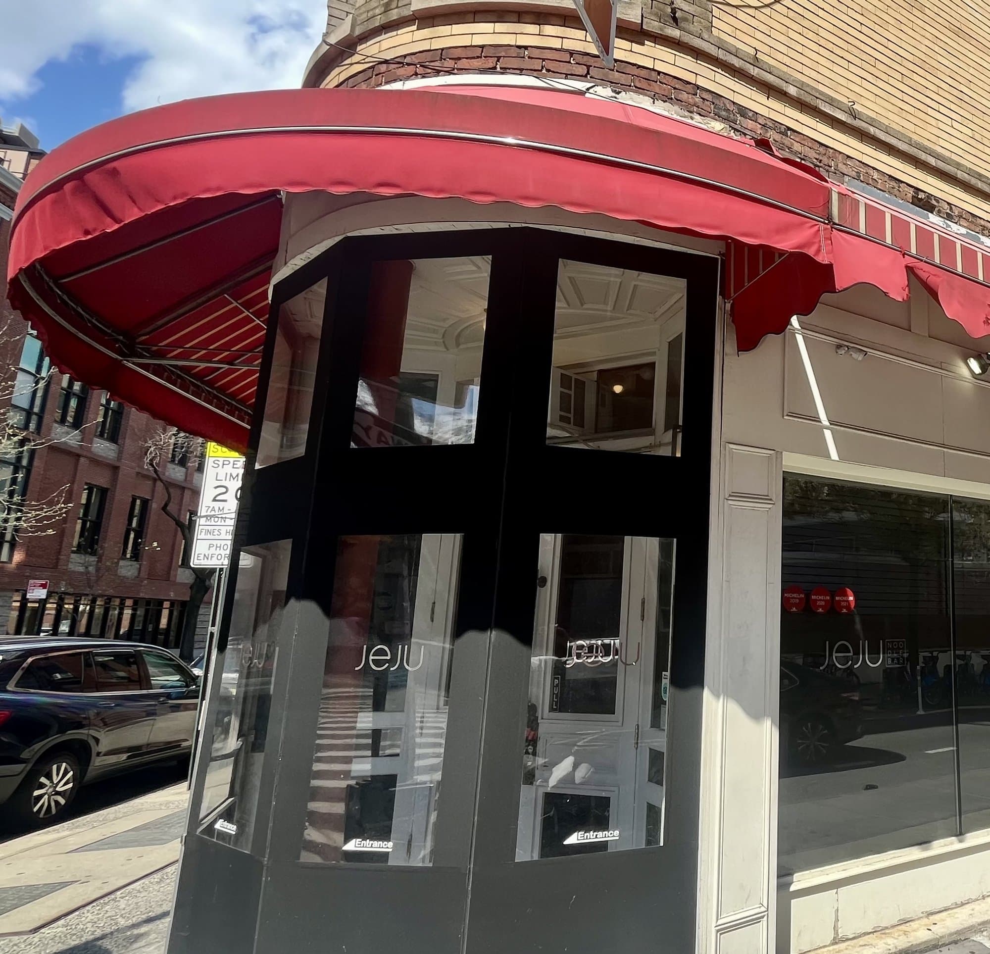 Corner entrance of Jeju Noodle Bar on Greenwich Street, with a red awning and the name lettered on the glass door