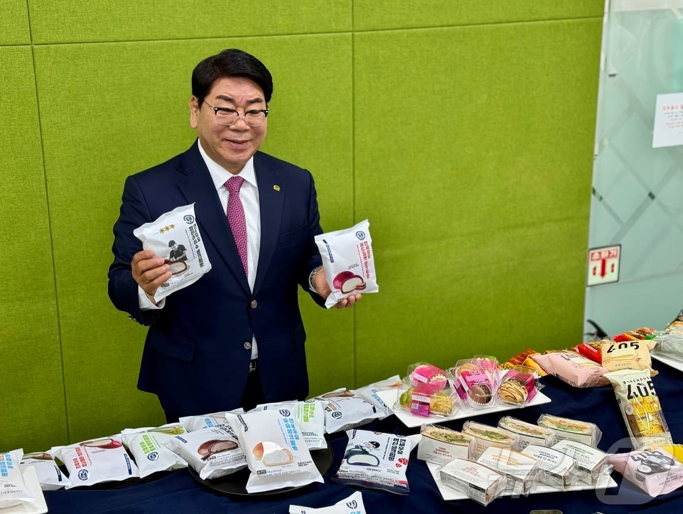 Kim Young-sik in a suit holding packaged cream breads behind a table of bakery products