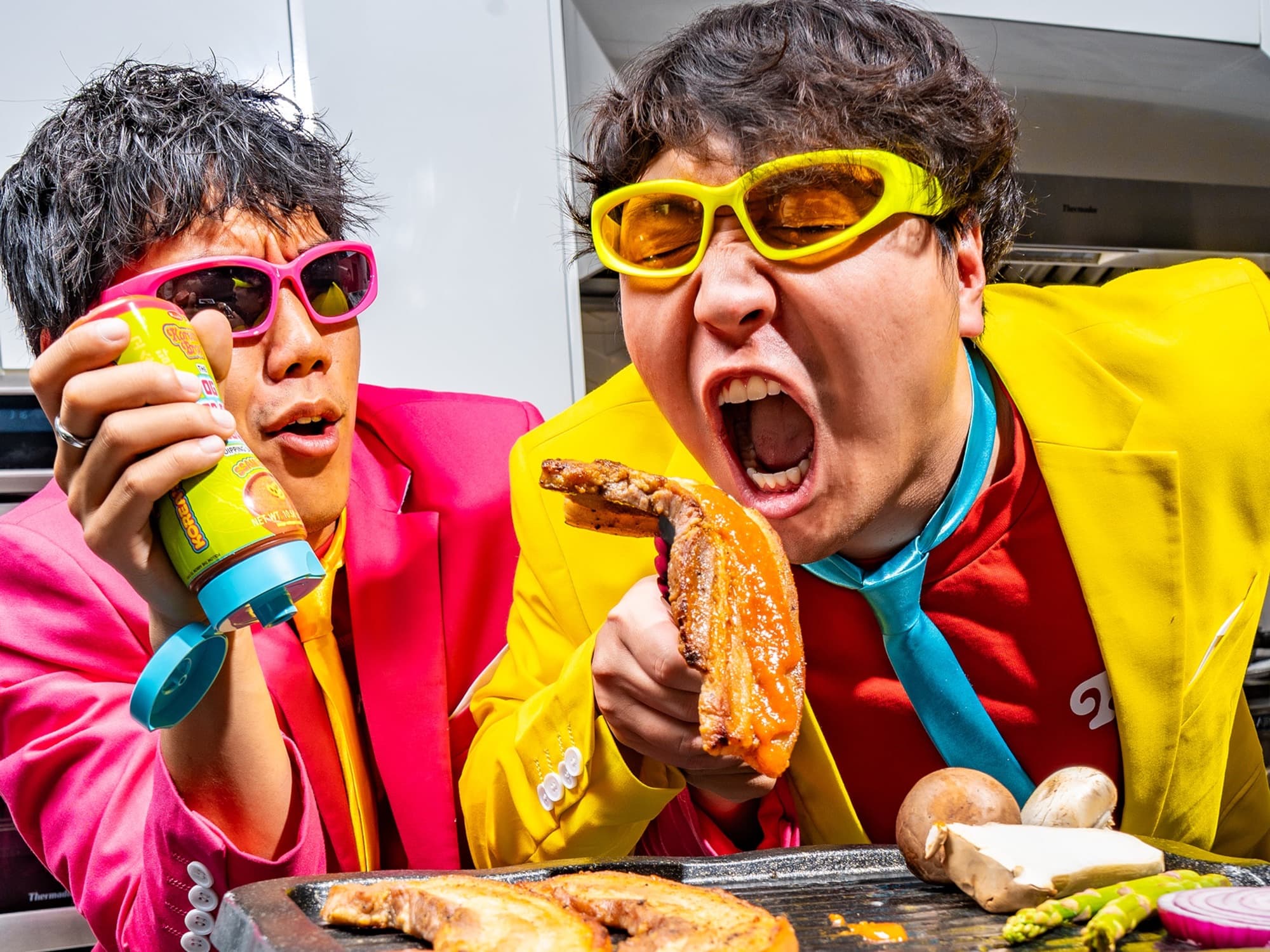 Korean Bros co-founders in bright suits sharing grilled pork belly and Korean BBQ sauce in a kitchen