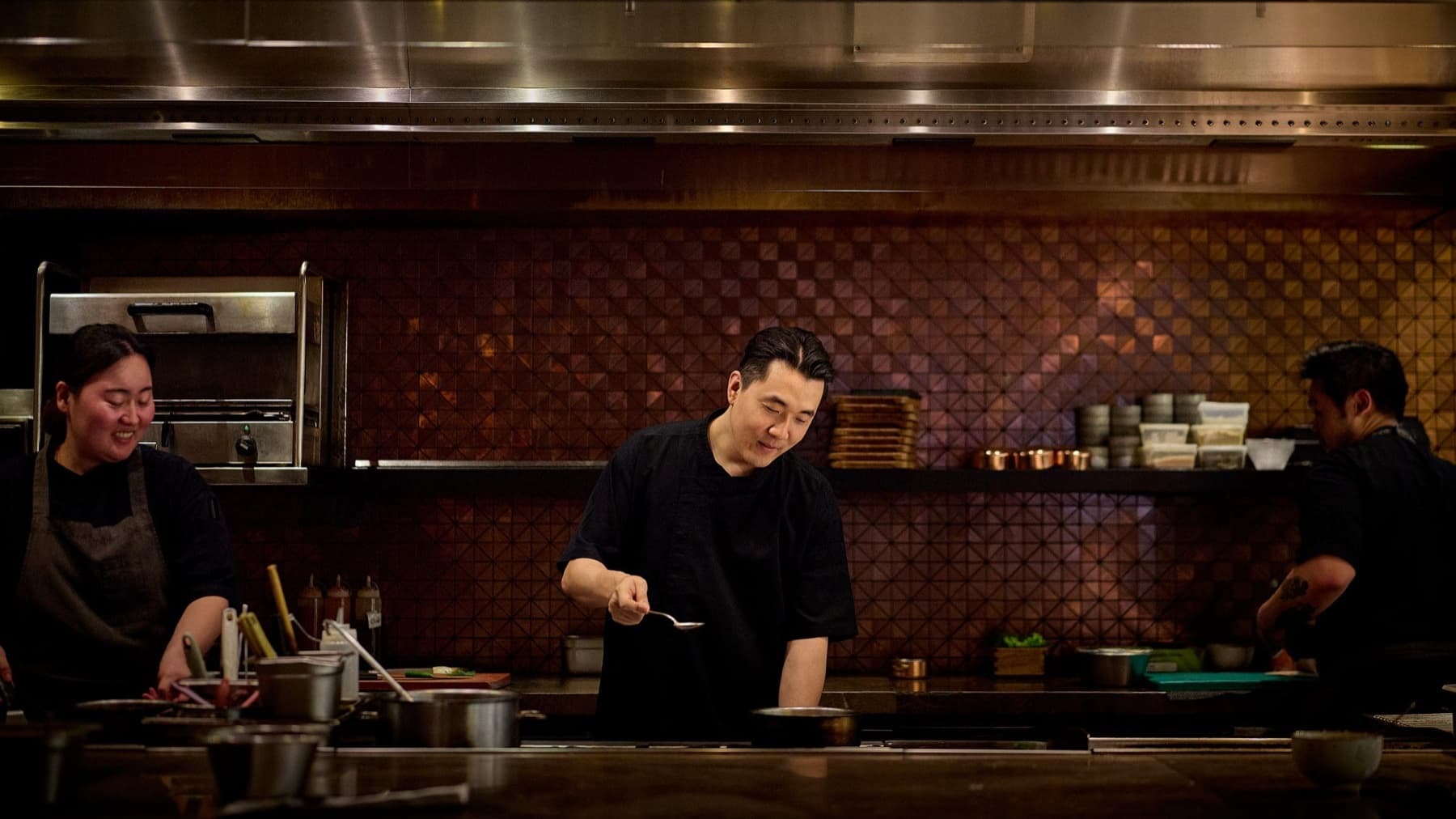 Chef Louis Han tasting from a spoon in a dark-tiled hotel kitchen with his team
