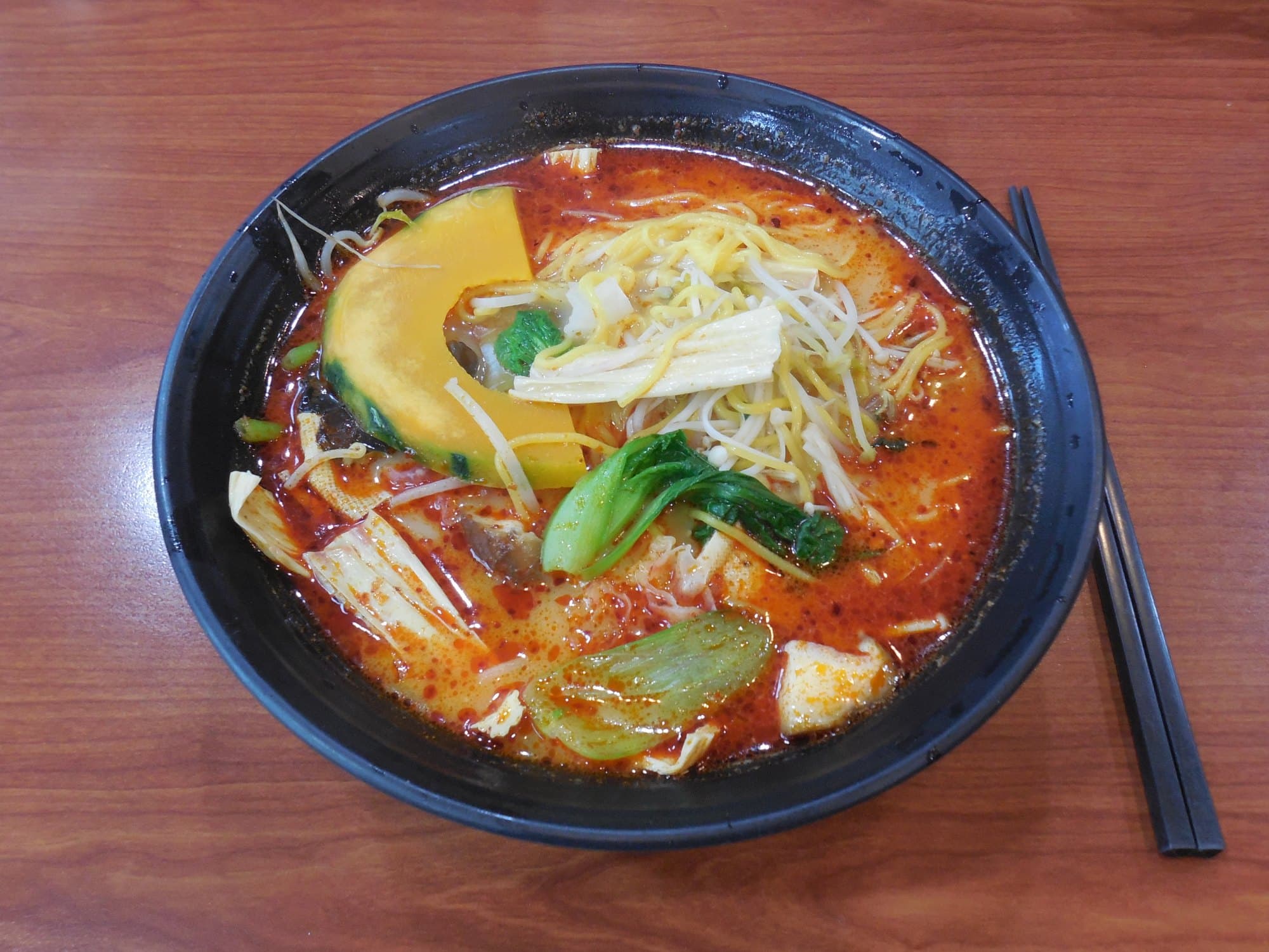 Overhead bowl of red malatang broth with squash, greens, and noodles on a wood table