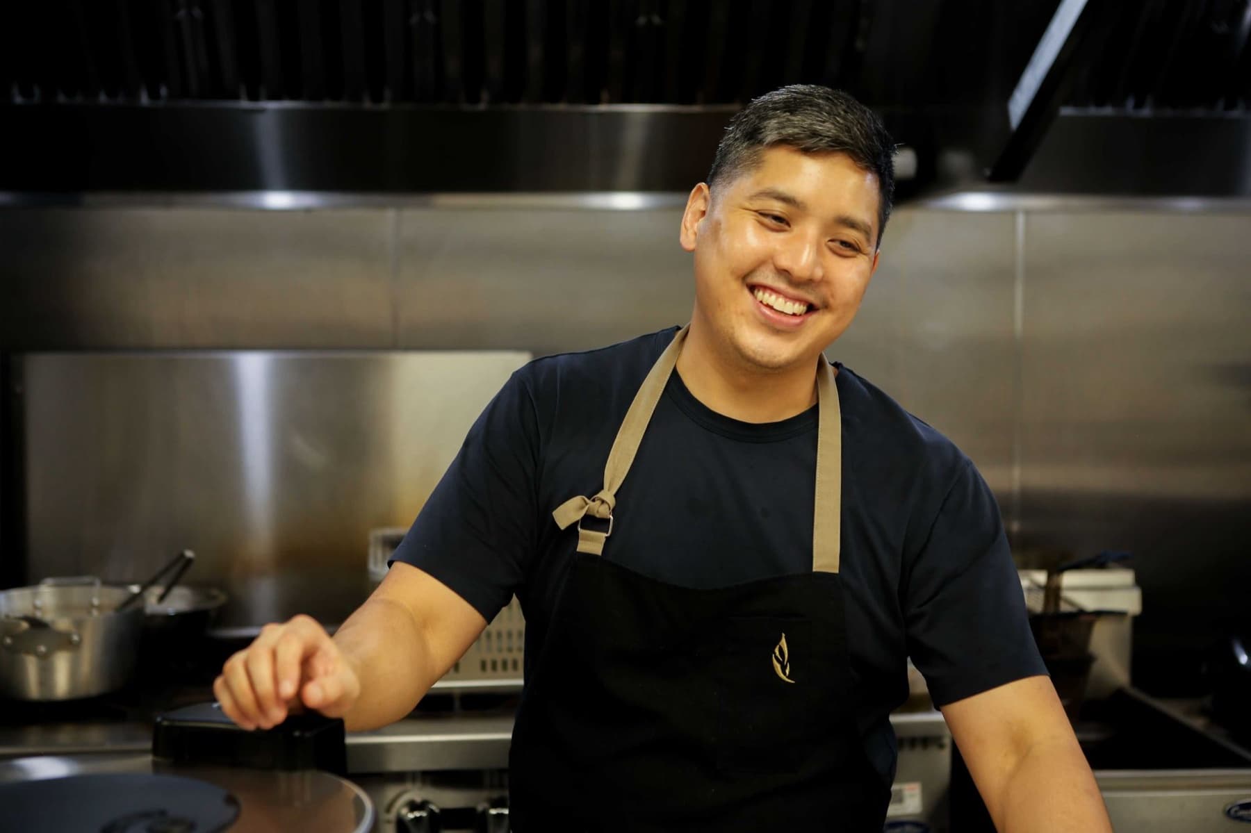 Chef Kris Hoang in a black apron smiling in a stainless-steel kitchen