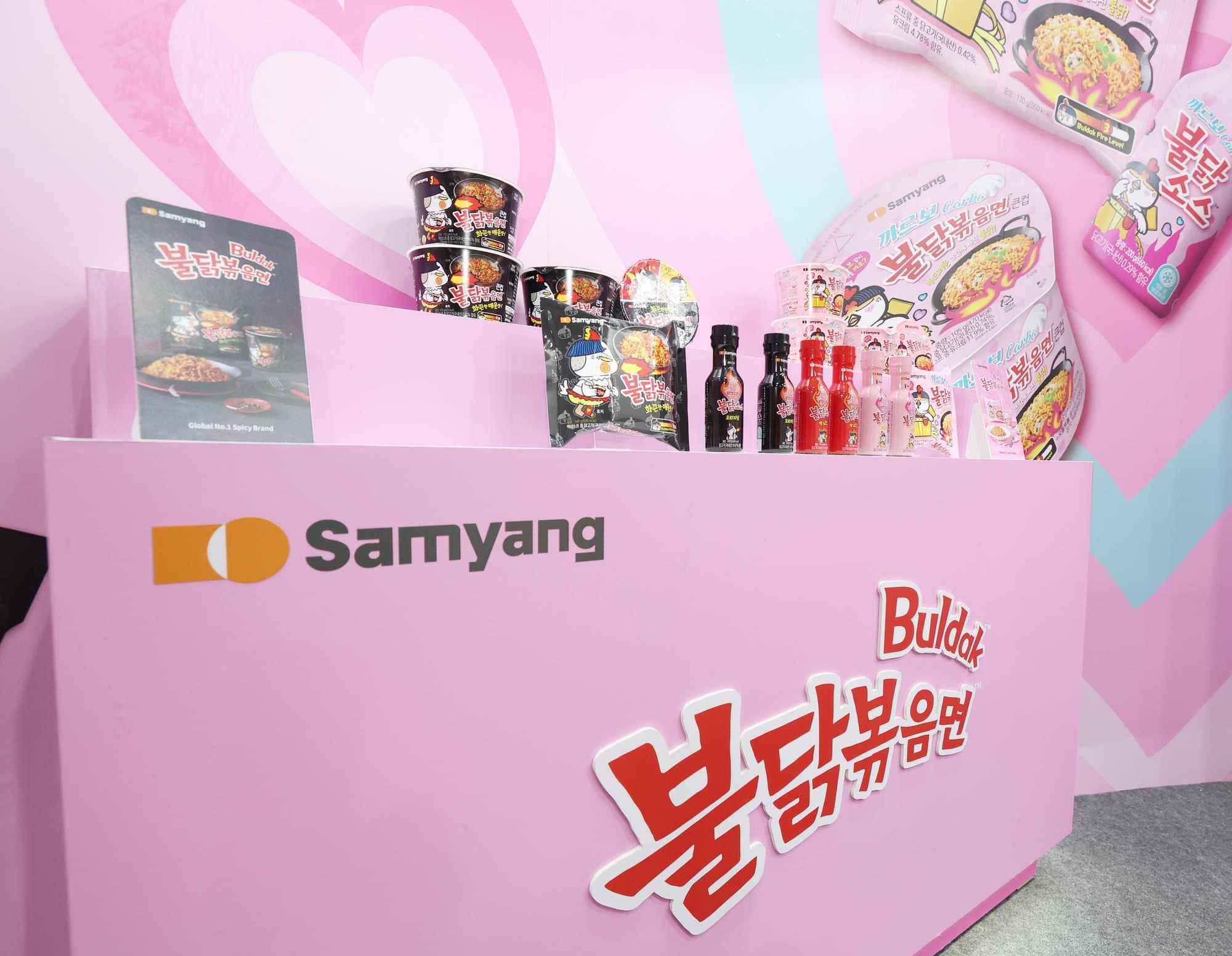 A pink Samyang booth displaying Buldak cups, packets, and sauce bottles under the brand name