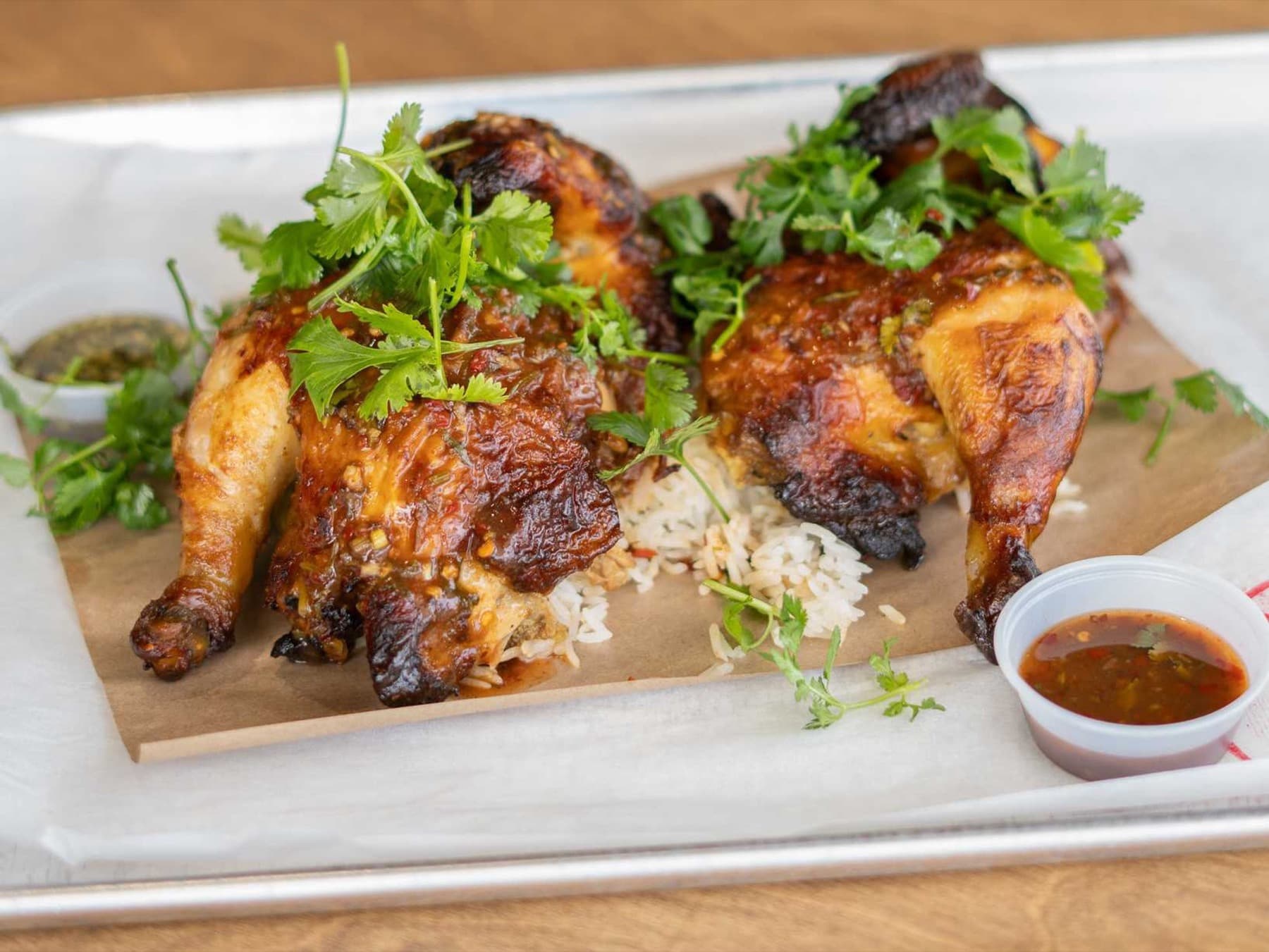 Thai-style rotisserie chicken halves on jasmine rice with cilantro and dipping sauces on a metal tray