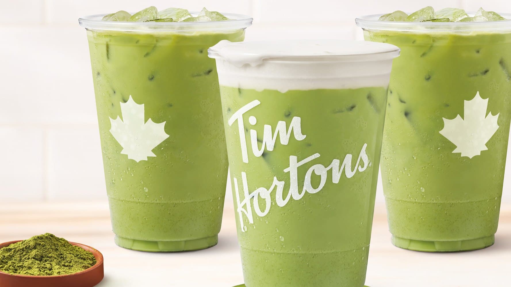 Three Tim Hortons iced matcha drinks on a wood counter beside a bowl of matcha powder