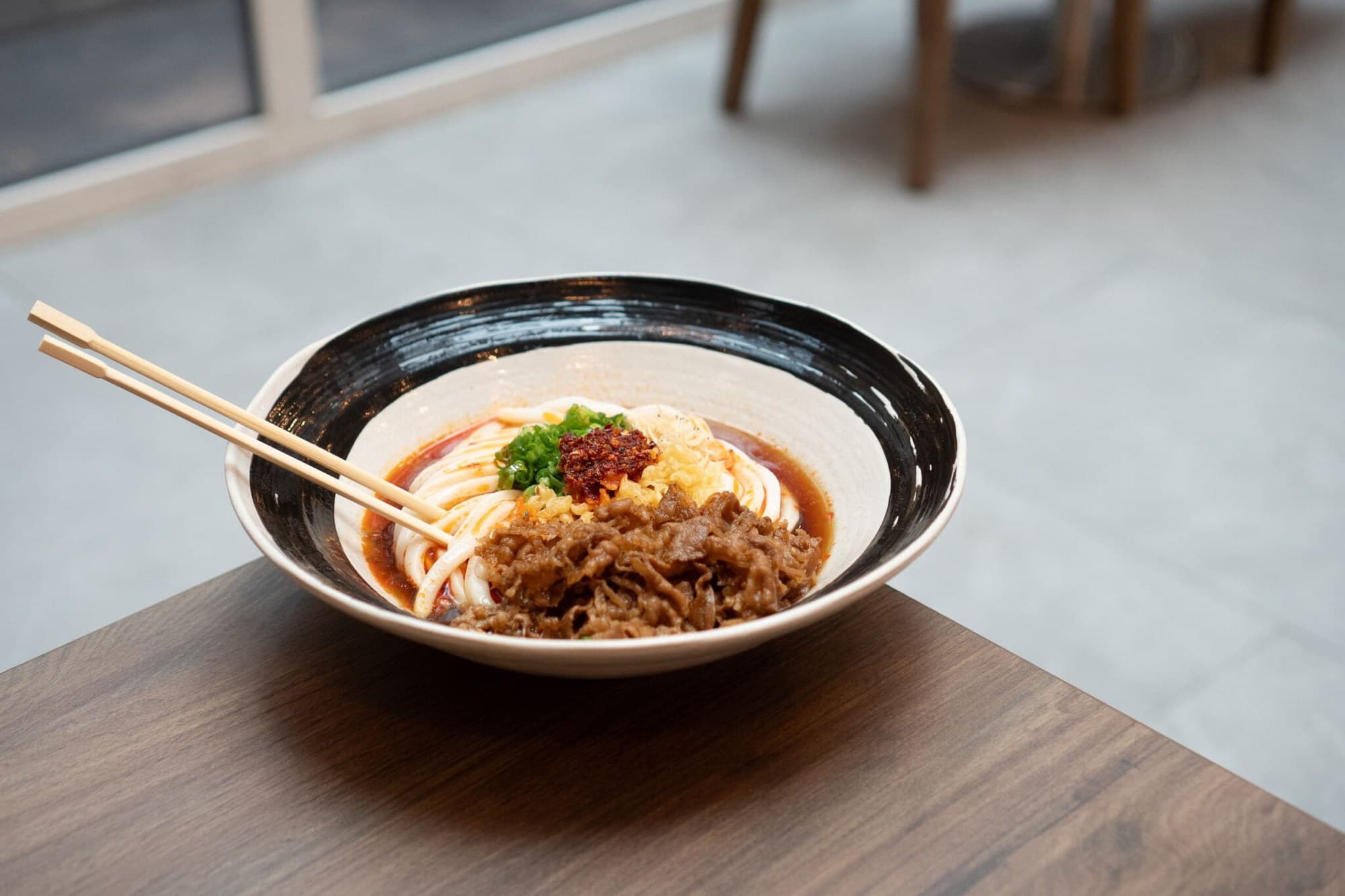 A wide bowl of spicy cold beef udon with chili oil, scallions, and tempura flakes at Yume Ga Arukara